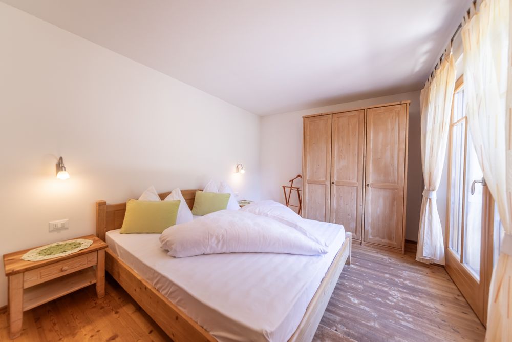 A cozy bedroom with a wooden double bed, a large wooden wardrobe, and light curtains next to a balcony door.