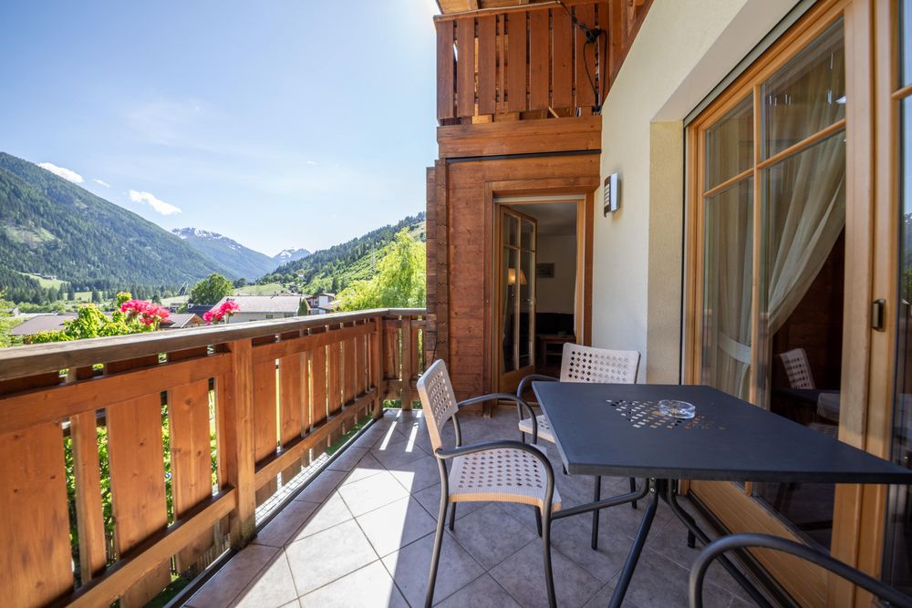 A sunny wooden balcony with seating furniture and a view of the Alps.